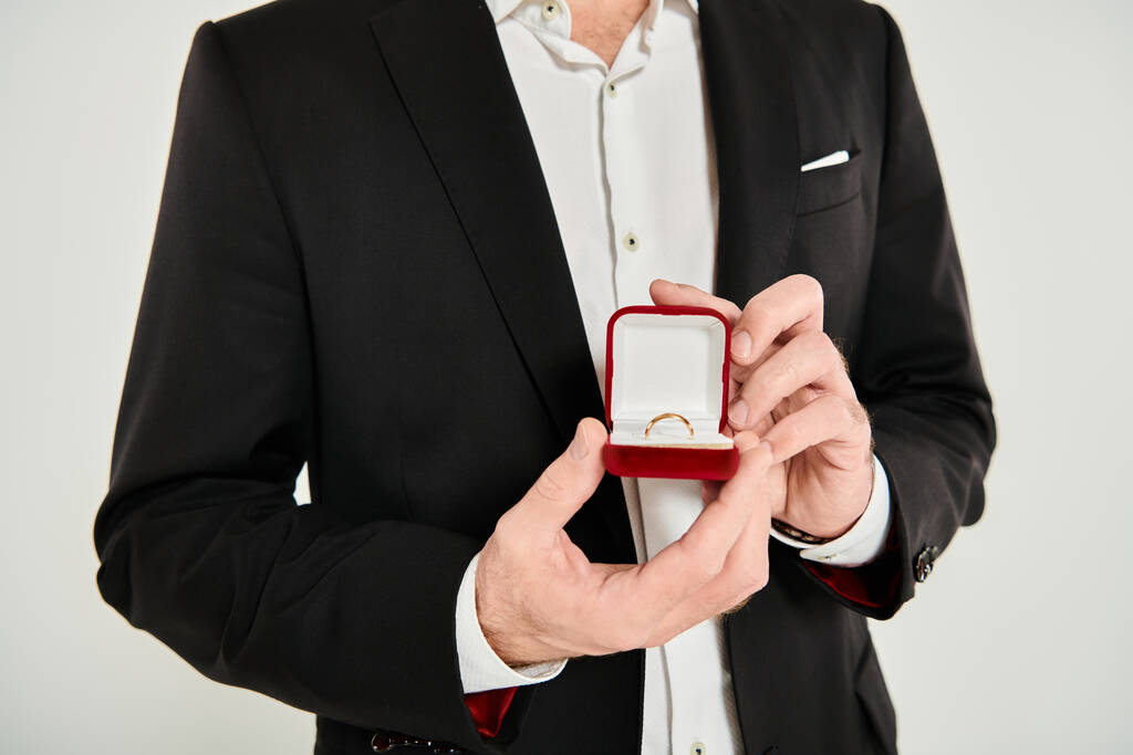 cropped άποψη του ανθρώπου σε μαύρο κοστούμι δείχνει κοσμήματα κουτί με δαχτυλίδι σε γκρι, Άγιος Βαλεντίνος έννοια - Φωτογραφία, εικόνα