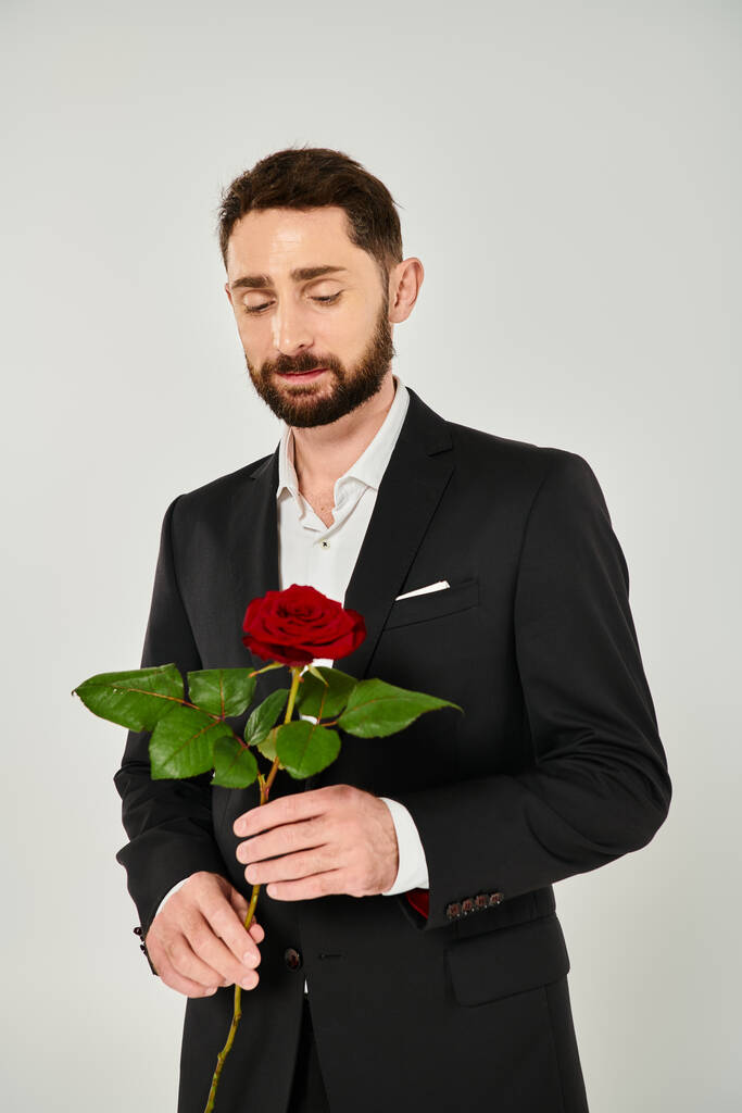 álmodozó szakállas férfi fekete elegáns öltöny kezében vörös rózsa szürke, Szent Valentin nap koncepció - Fotó, kép