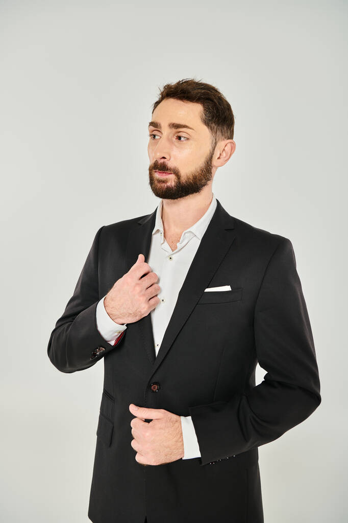 стомлений елегантний бородатий бізнесмен в чорному костюмі, стоячи і дивлячись на сірий фон - Фото, зображення
