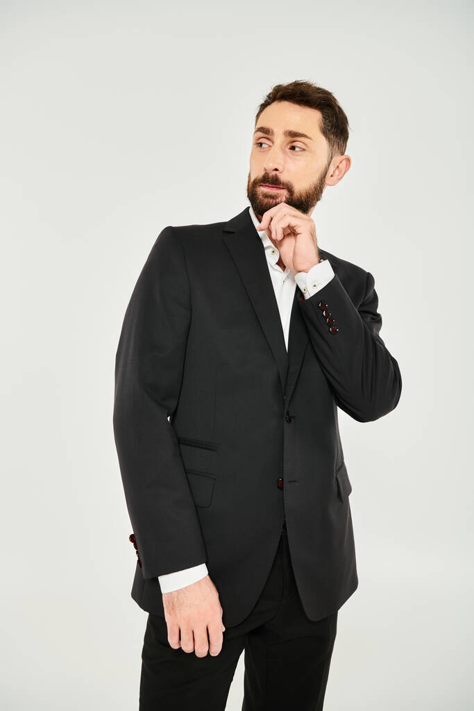 pensativo hombre de negocios elegante en traje negro tocando la barba y mirando hacia otro lado en el fondo gris - Foto, imagen