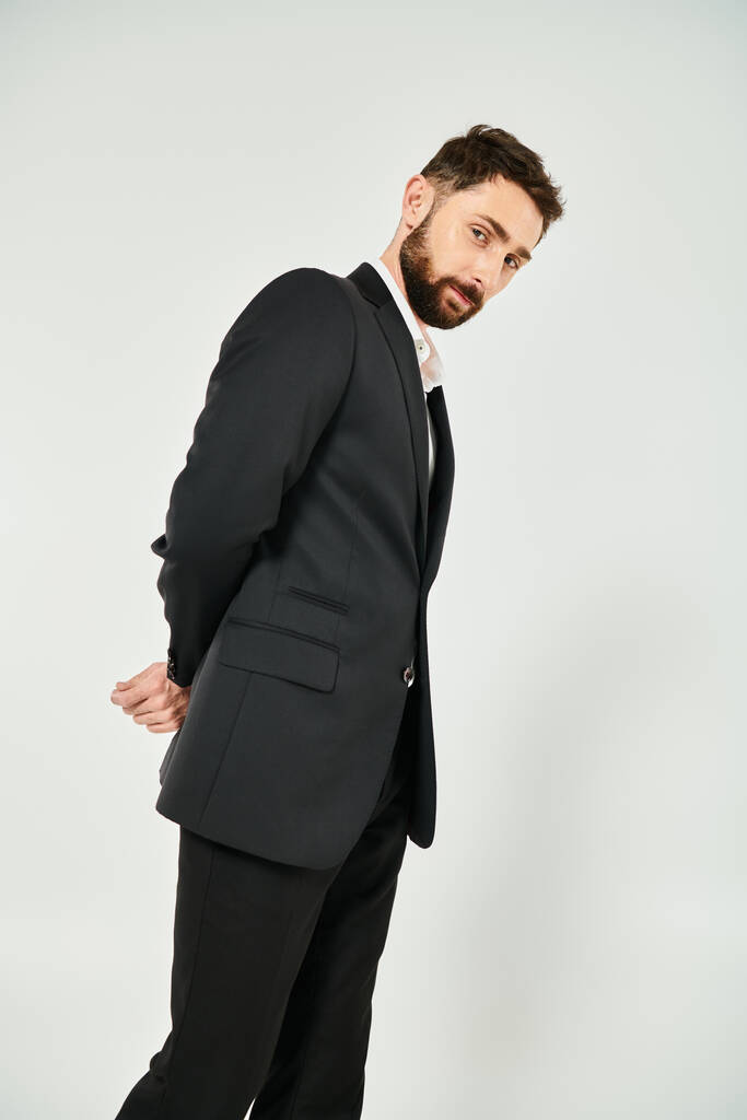 positive and elegant bearded businessman with hands behind back looking at camera on grey backdrop - Photo, Image