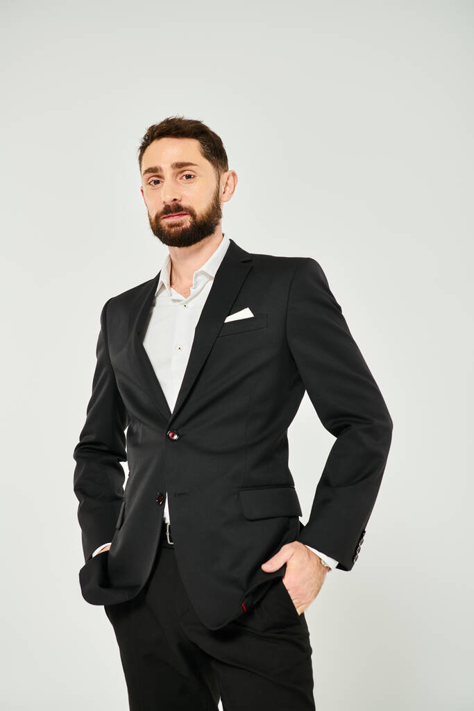 confident bearded businessman with hands in pockets looking at camera while posing on grey - Photo, Image