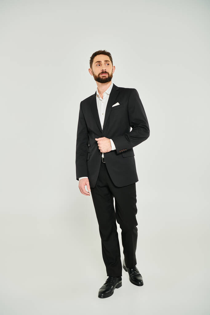 rico y exitoso hombre de negocios en traje negro de pie y mirando hacia otro lado en gris, longitud completa - Foto, imagen