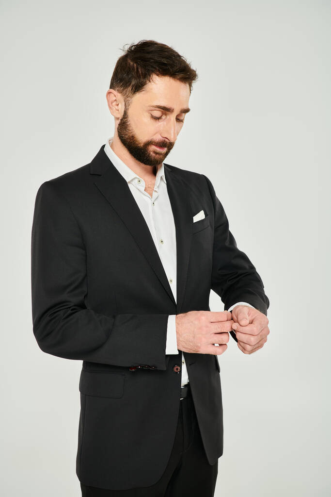 hombre de negocios bien vestido ajustando la manga de traje elegante negro mientras está de pie sobre fondo gris - Foto, imagen
