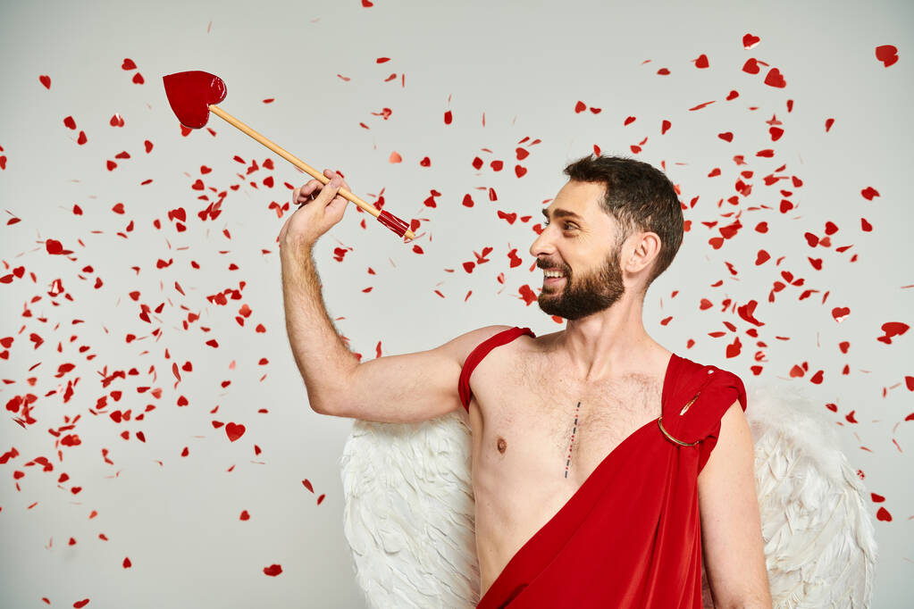alegre barbudo hombre Cupido con flecha en forma de corazón bajo confeti rojo en gris, día de San Valentín - Foto, imagen