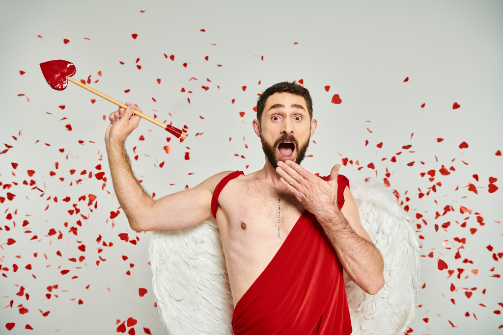 amazed bearded cupid man with arrow covering open mouth with hand under red heart-shaped confetti - Photo, Image