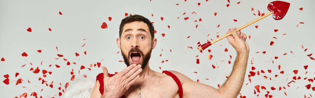 astonished cupid man holding arrow and covering open mouth with hand under red confetti, banner - Photo, Image