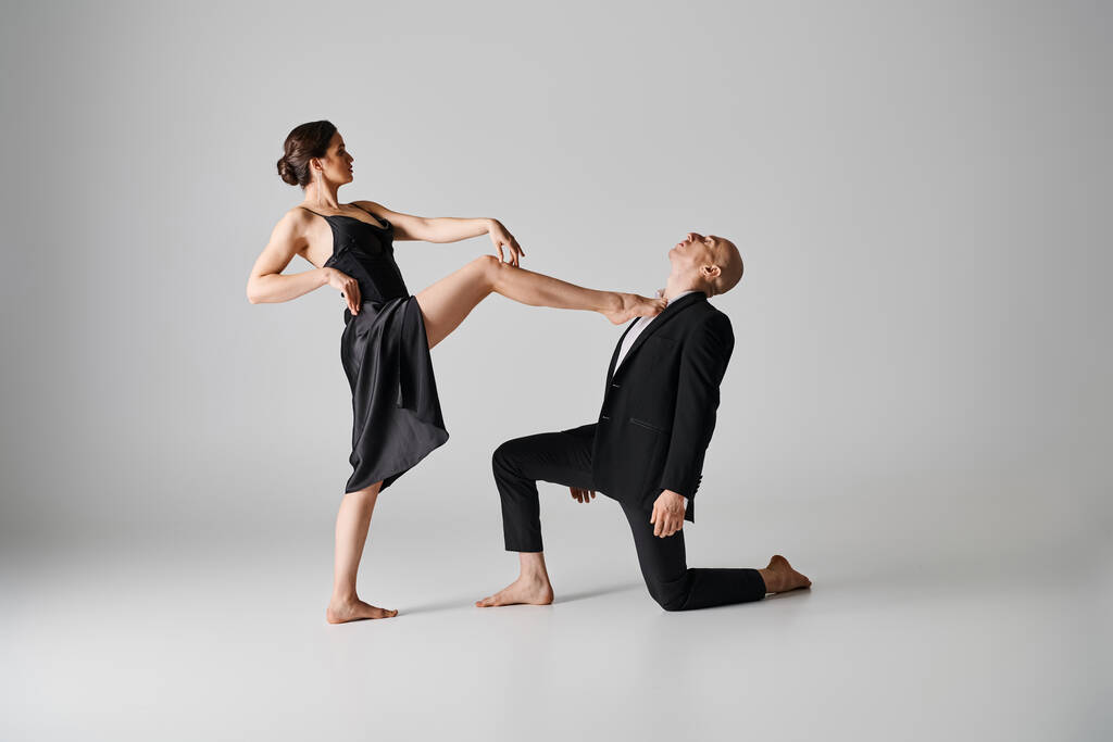 Siyah elbiseli genç çıplak ayaklı kadın gri arka plandaki adamla tutkulu bir dans gösterisi yapıyor. - Fotoğraf, Görsel