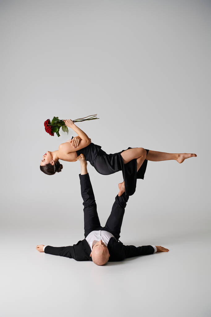 ευέλικτη γυναίκα με μαύρη ενδυμασία που κρατάει κόκκινα τριαντάφυλλα και ισορροπεί στα πόδια του παρτενέρ που χορεύει στο γκρι - Φωτογραφία, εικόνα