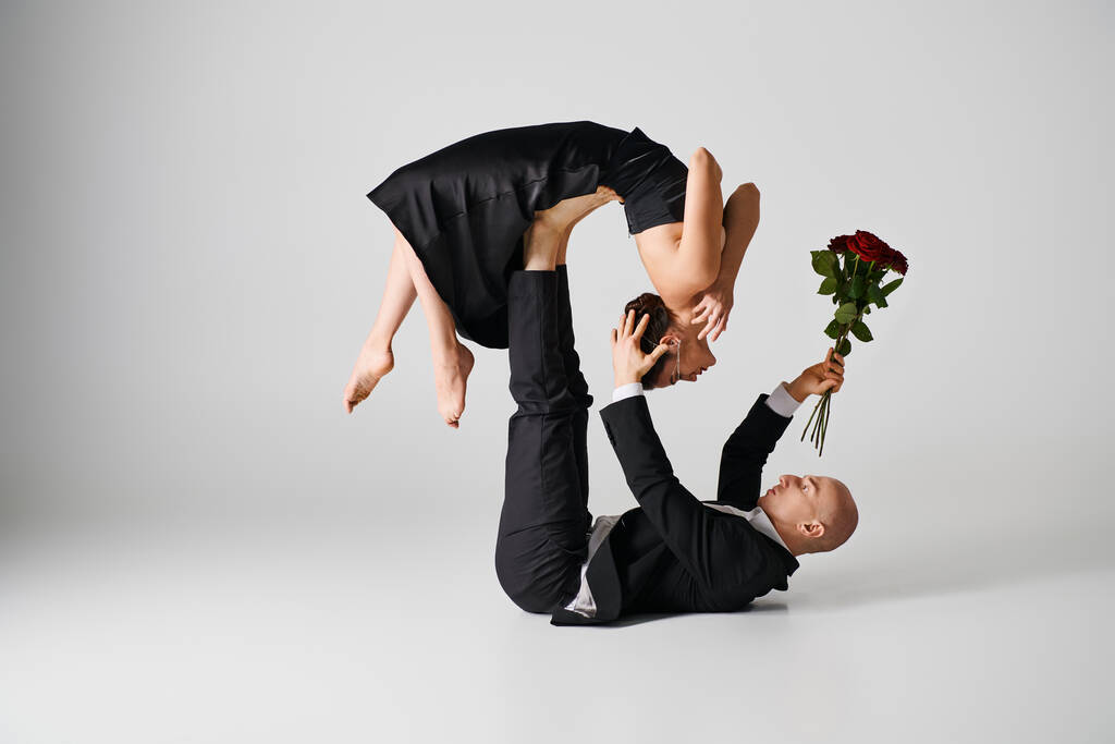 νεαρή ευέλικτη γυναίκα με μαύρη ενδυμασία που ισορροπεί στα πόδια του συντρόφου που χορεύει κρατώντας κόκκινα τριαντάφυλλα σε γκρι - Φωτογραφία, εικόνα