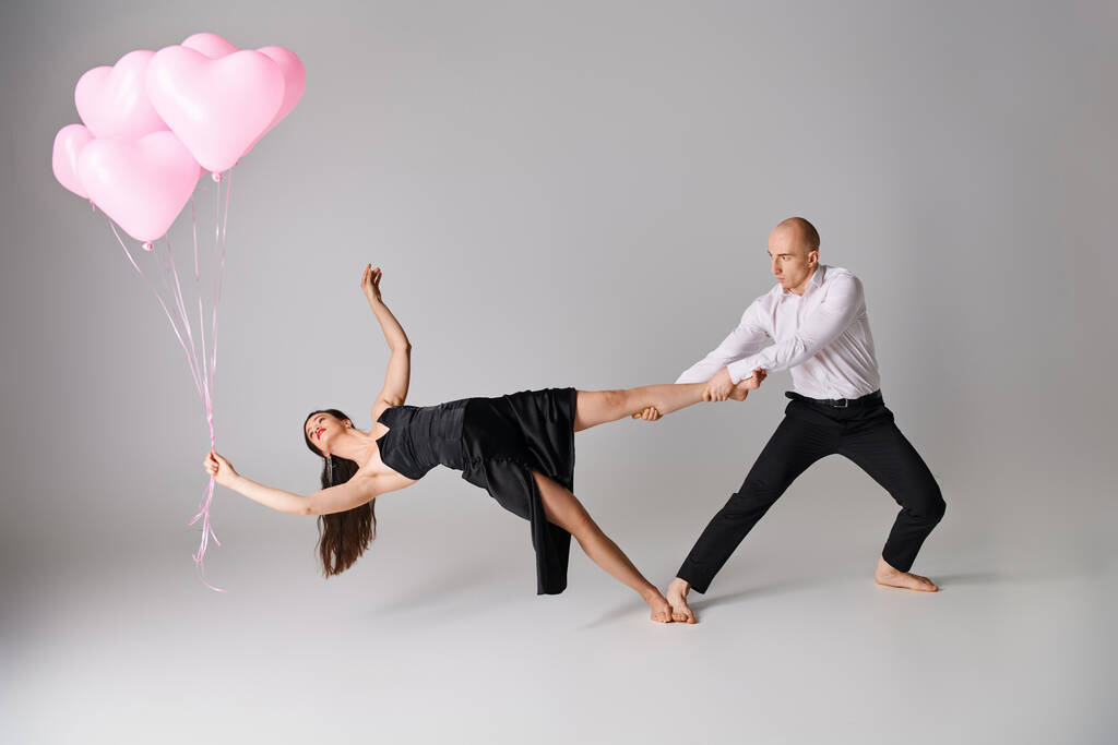 elastyczne młoda kobieta w czarny sukienka balansowanie na jej nagie nogi z balony w pobliżu mężczyzna na szary - Zdjęcie, obraz