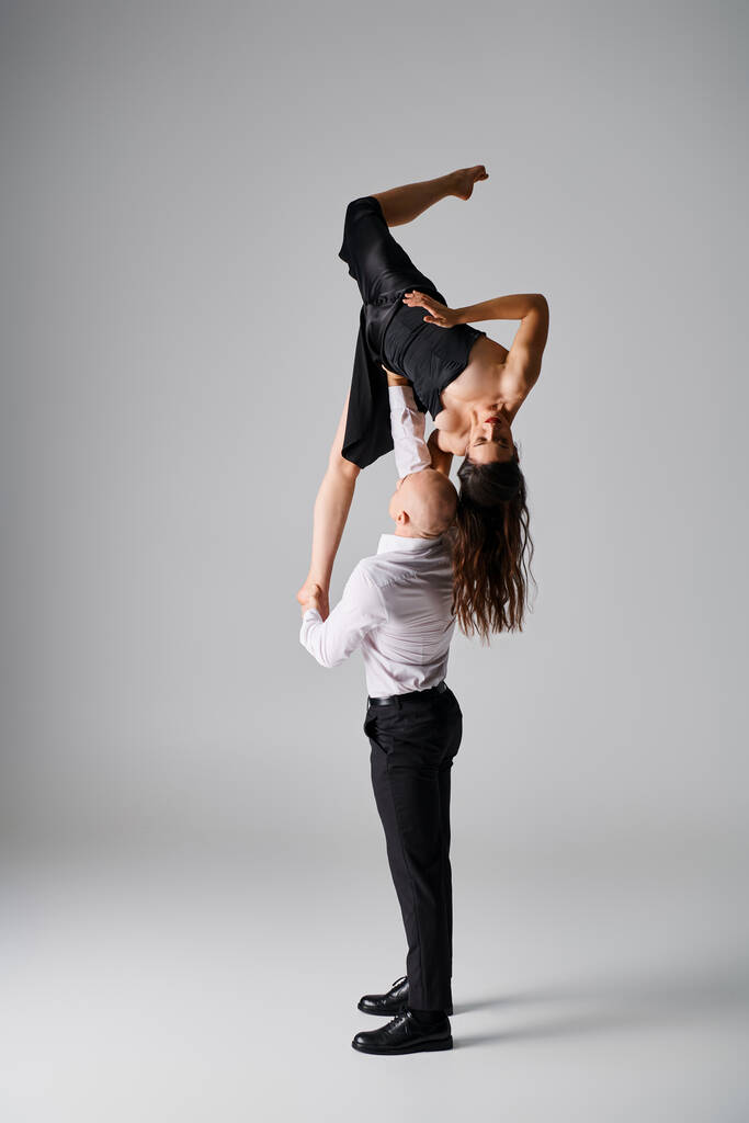 сильный мужчина поднимает брюнетку молодая женщина в черном платье во время исполнения танца на сером фоне - Фото, изображение