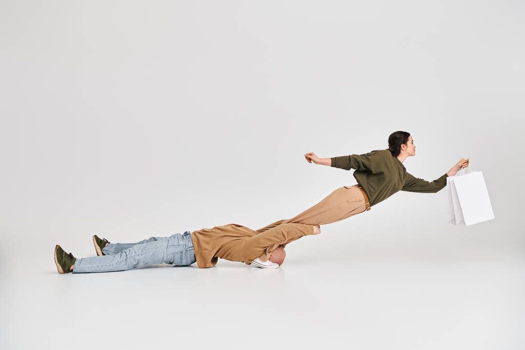acrobatic woman holding shopping bag and balancing in air with support of man in studio on grey - Photo, Image