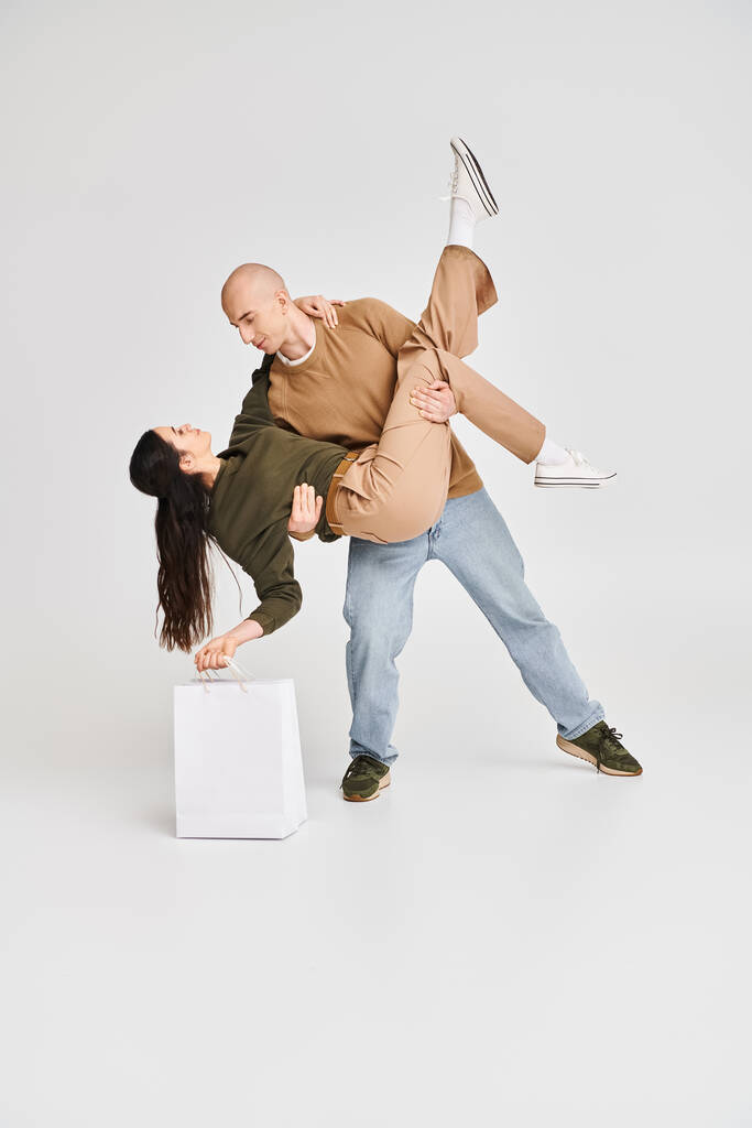 man in casual attire holding in arms beautiful brunette woman with shopping bag on grey backdrop - Photo, Image
