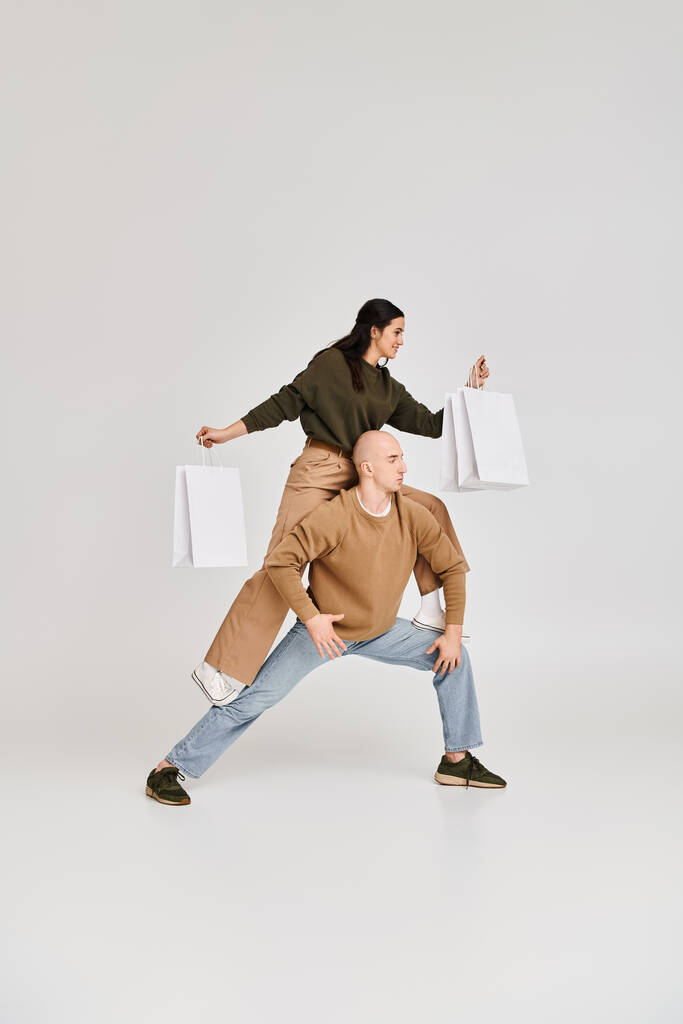 acrobatic performance of young couple, woman with shopping bags balancing on body of man on grey - Photo, Image