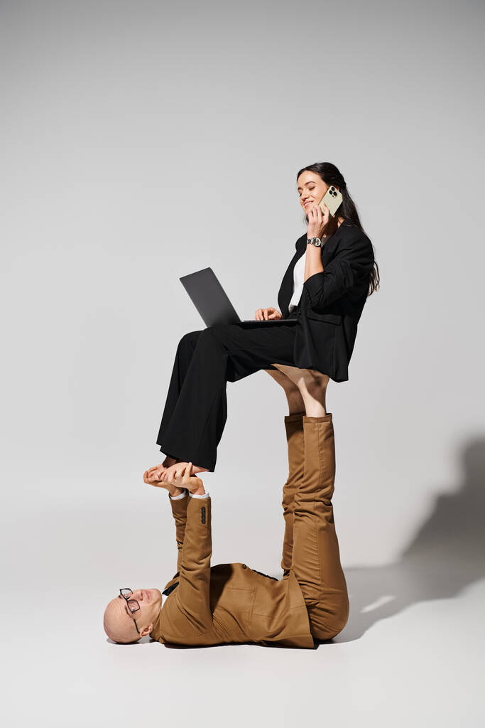 ευτυχισμένη γυναίκα στην επιχείρηση ενδυμασία με φορητό υπολογιστή έχοντας τηλεφωνική κλήση και εξισορρόπηση στα πόδια του άνδρα σε γκρι - Φωτογραφία, εικόνα