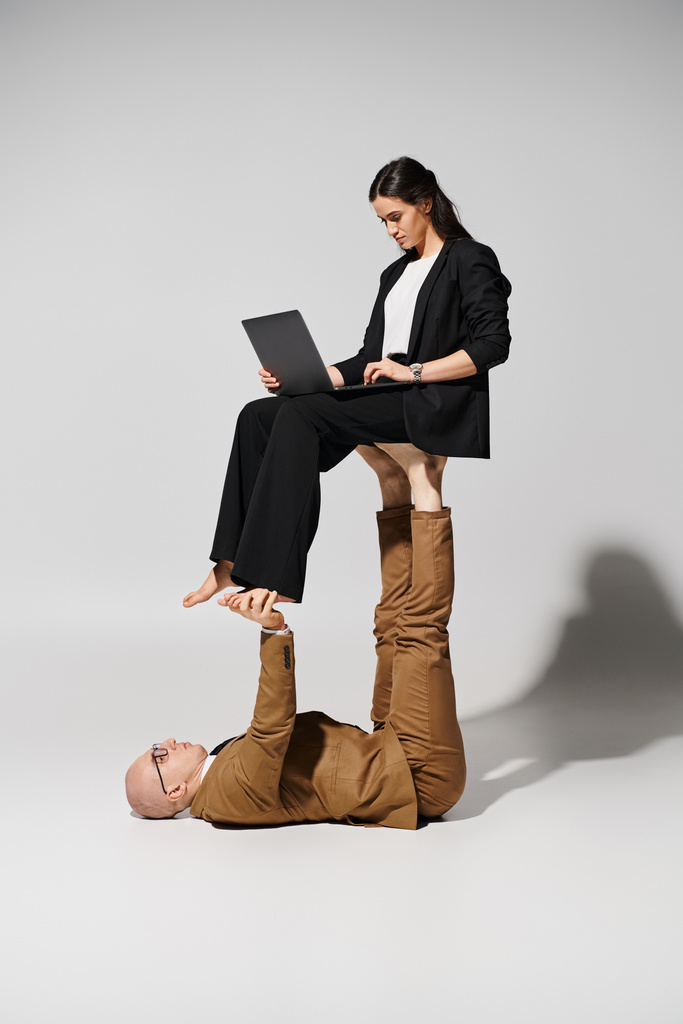 Geschäftsfrau in Anzug mit Laptop balanciert auf Füßen eines Mannes im Anzug auf grau, ein paar Akrobaten - Foto, Bild
