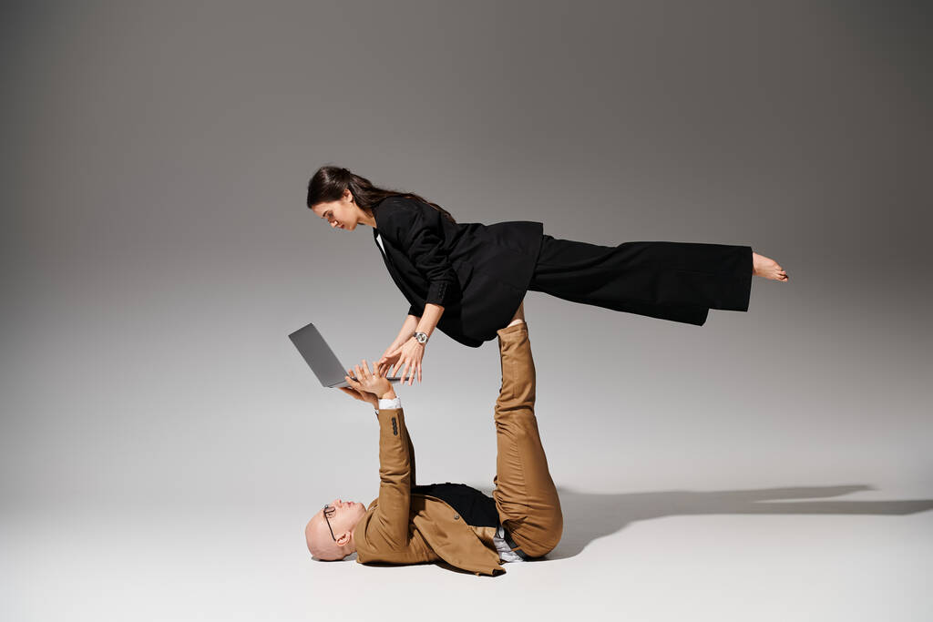 Kobieta w biznesie z laptopem balansującym na nogach mężczyzny w garniturze na szarym, para akrobatów - Zdjęcie, obraz