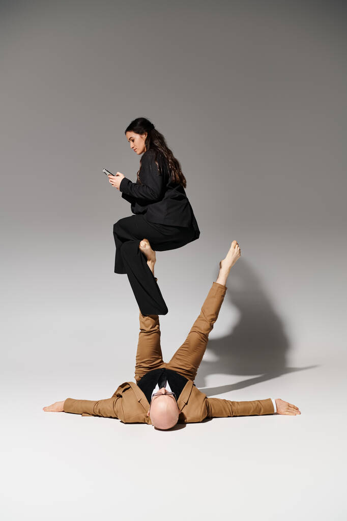 businesswoman ze smartfonem siedzi na nogach mężczyzny w garniturze na szarości, para akrobatów - Zdjęcie, obraz