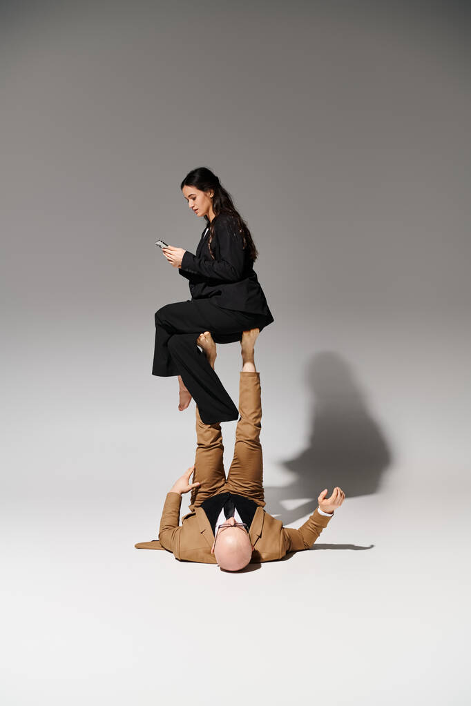 junge Geschäftsfrau mit Smartphone balanciert auf den Füßen eines Mannes im Anzug auf grau, ein paar Akrobaten - Foto, Bild