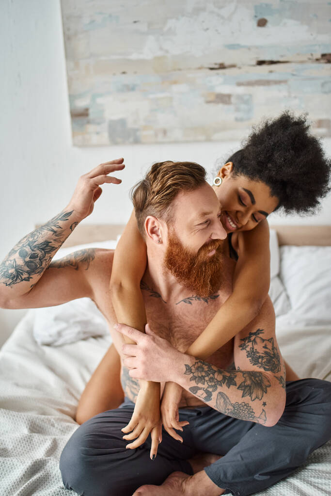 gelukkig divers paar, krullend Afrikaans amerikaans vrouw omarmen haar getatoeëerde vriend in slaapkamer - Foto, afbeelding
