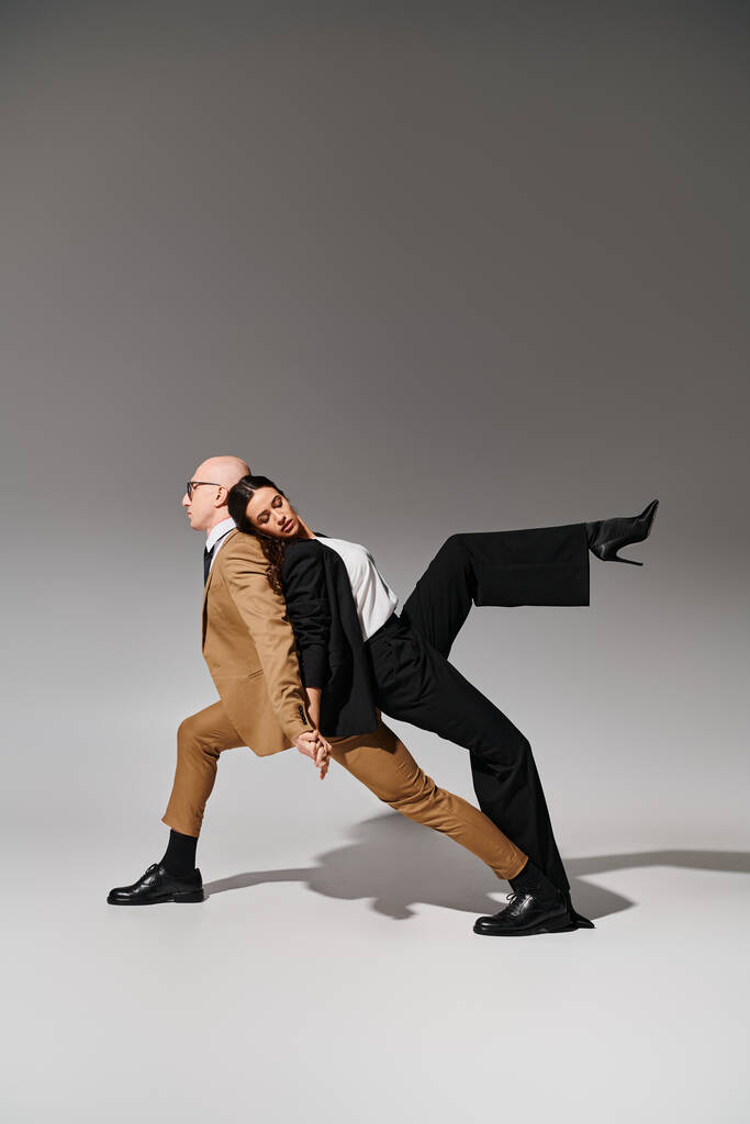 danspartners in gecoördineerde beweging, vrouw in pak leunend op rug van man in zakelijke kleding op grijs - Foto, afbeelding