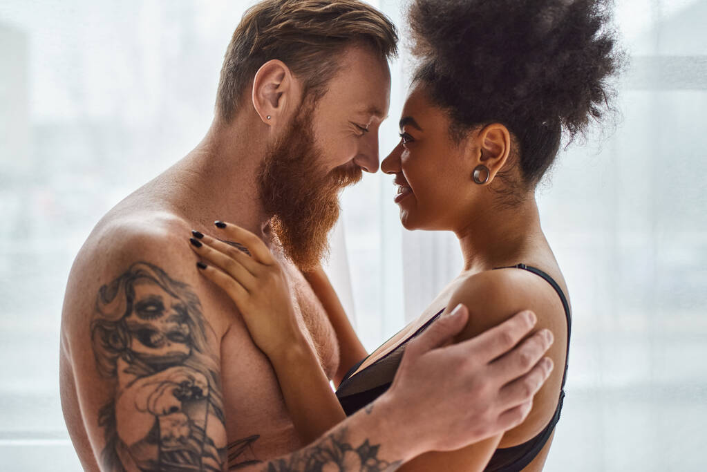 happy bearded and tattooed man without shirt hugging his african american girlfriend near curtain - Photo, Image