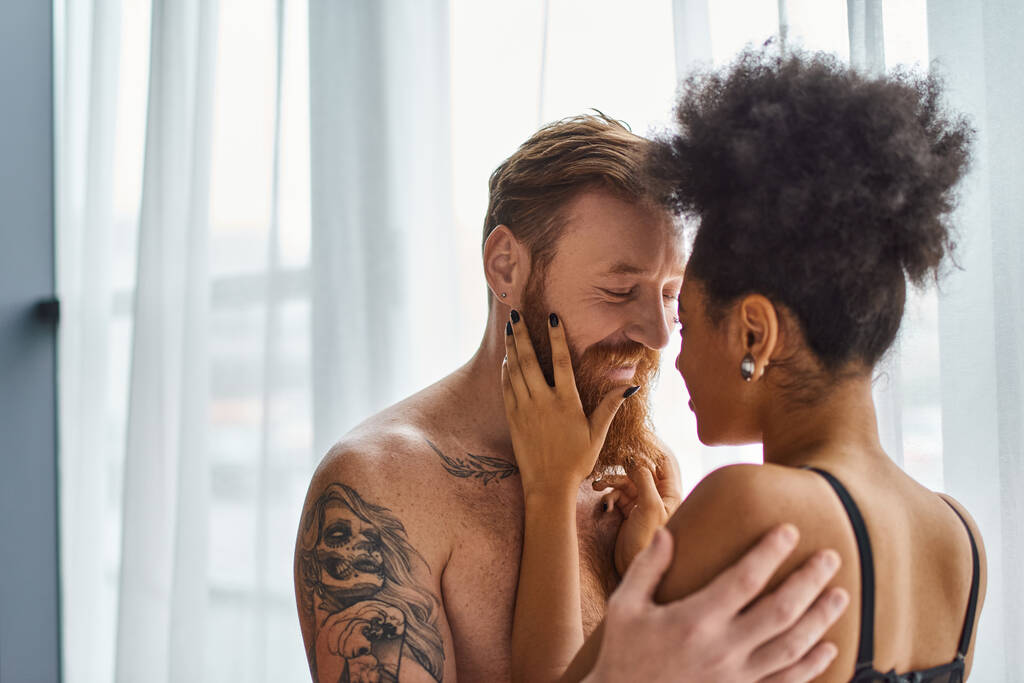 heureux homme barbu et tatoué sans chemise étreignant sa petite amie afro-américaine près du rideau - Photo, image