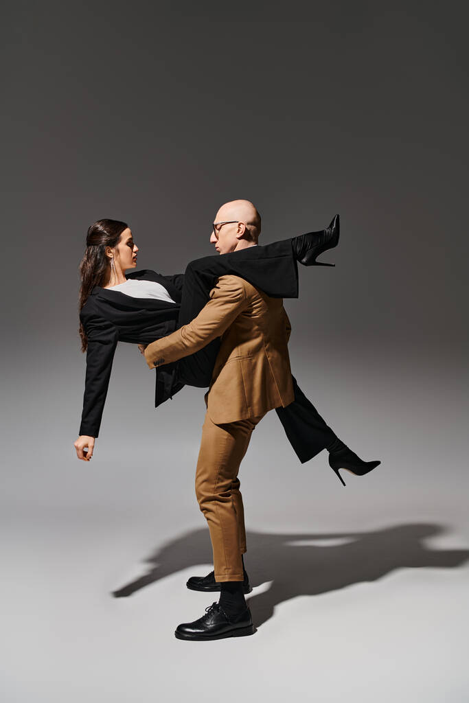 Gözlüklü ve takım elbiseli bir adam yüksek topuklu esmer bir kadını kaldırıyor ve gri arka planda resmi giyiniyor. - Fotoğraf, Görsel