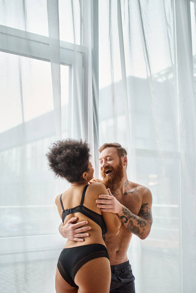 hombre excitado y tatuado sin camisa abrazando a su novia afroamericana feliz cerca de la cortina - Foto, Imagen