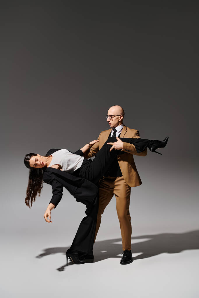 グラスとスーツの男は,灰色の背景でダンス中にハイヒールでブルネット女性の脚をサポート - 写真・画像