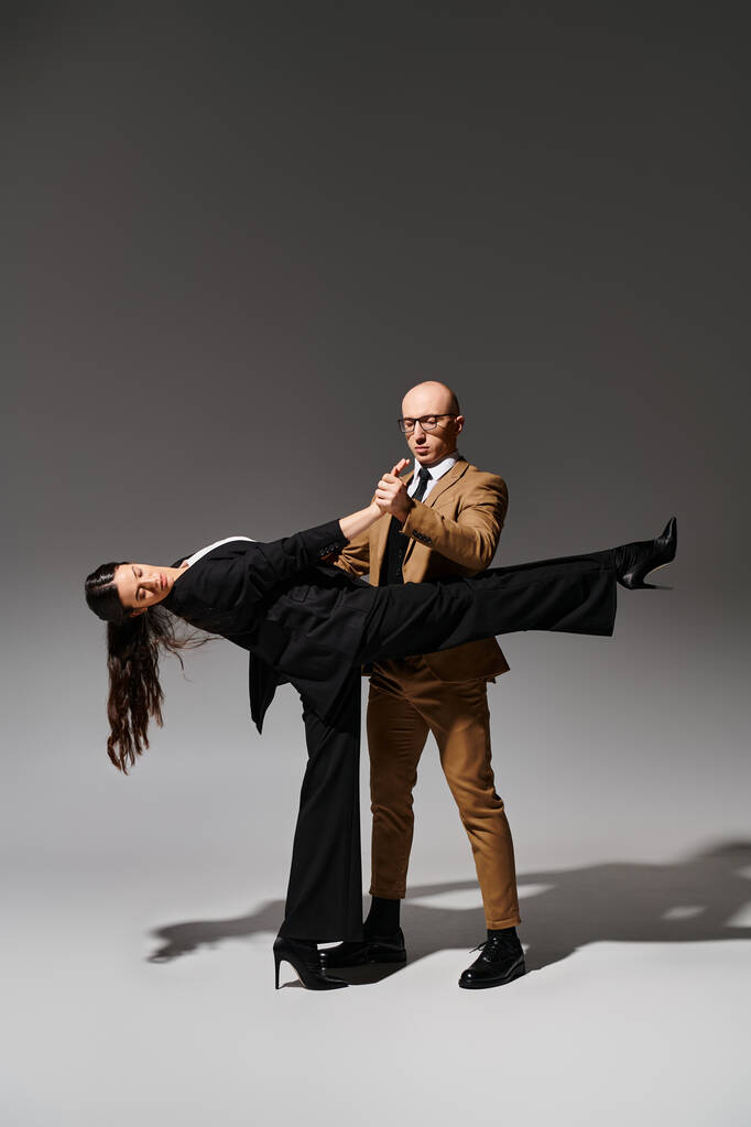mężczyzna w okularach i garniturze trzymając rękę brunetka kobieta w wysokich obcasach podczas tańca na szarym tle - Zdjęcie, obraz