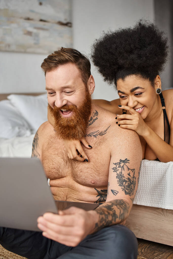 Mutlu dövmeli adam ve Afro-Amerikan kadın dizüstü bilgisayarda komedi filmi izliyorlar, çok kültürlü çift. - Fotoğraf, Görsel