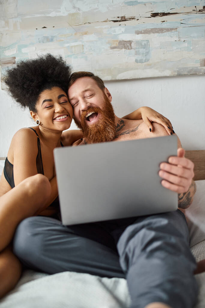 счастливая африканская американка в нижнем белье с помощью ноутбука и лежа на кровати рядом с татуированным мужчиной, фильм - Фото, изображение