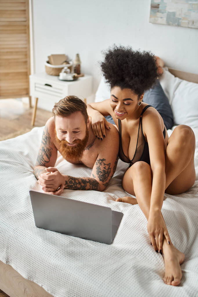 pareja multicultural viendo película de comedia en la cama, mujer afroamericana y hombre tatuado barbudo - Foto, Imagen