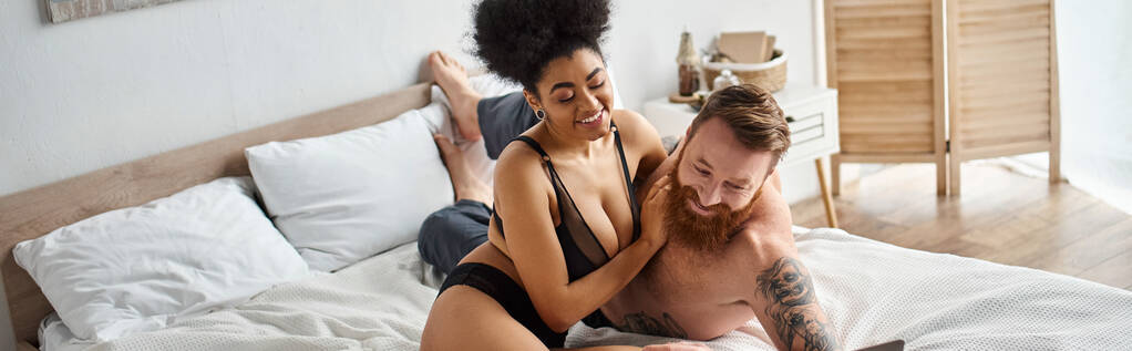 счастливый и татуированный мужчина без рубашки смеется в постели с кудрявой африканской американской девушкой, баннер - Фото, изображение