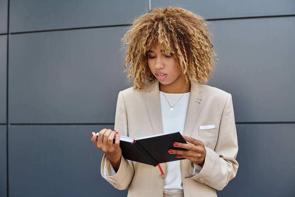 zamyślony afrykański amerykański bizneswoman w formalnym stroju patrząc na jej notatnik w pobliżu biurowca - Zdjęcie, obraz