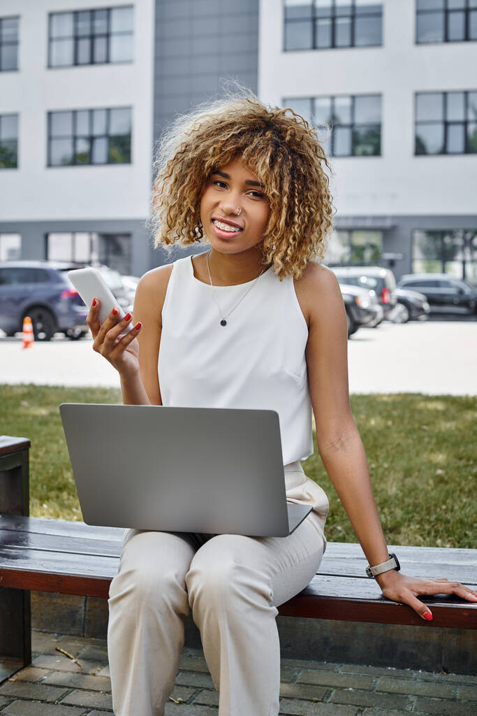 Χαρούμενη Αφροαμερικανή ελεύθερη επαγγελματίας με smartphone που δουλεύει στο laptop της σε παγκάκι σε πάρκο - Φωτογραφία, εικόνα