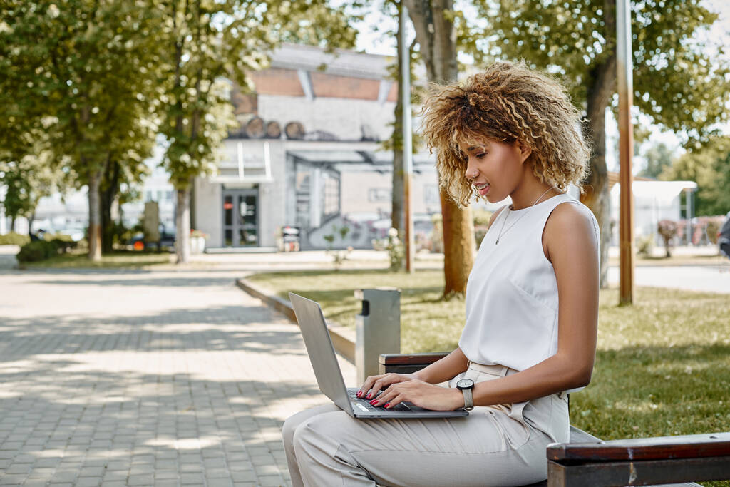 Geconcentreerd krullend Afrikaans Amerikaans freelancer werken op haar laptop op bank in een park instelling - Foto, afbeelding