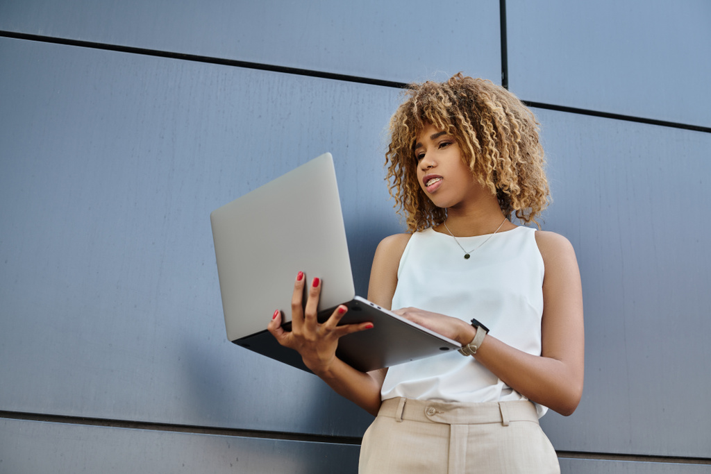 curly african american freelancer with braces standing with her laptop near grey wall of building - Photo, Image