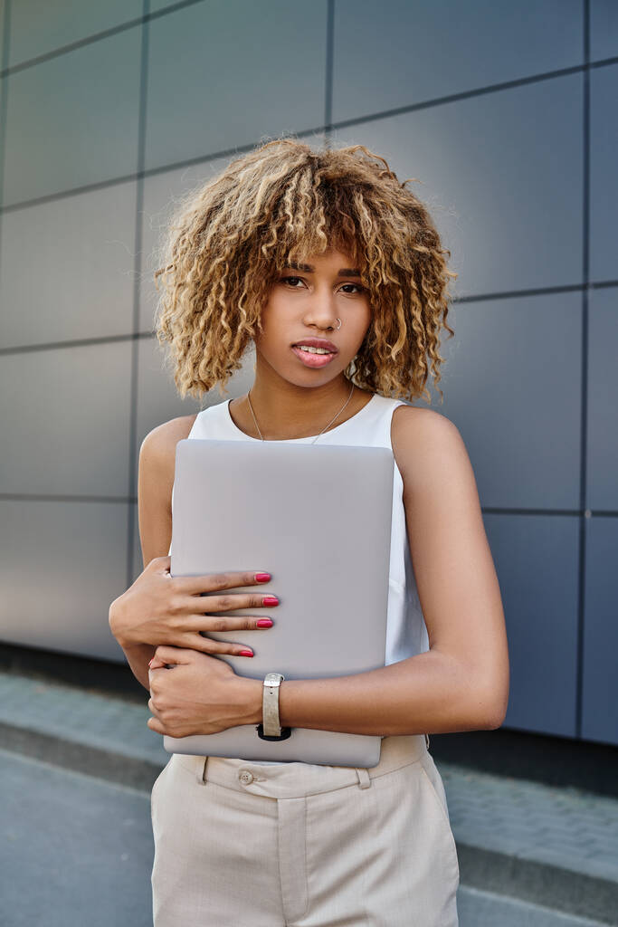 lockige afrikanisch-amerikanische Geschäftsfrau mit Zahnspange steht mit ihrem Laptop in der Nähe eines Bürogebäudes - Foto, Bild
