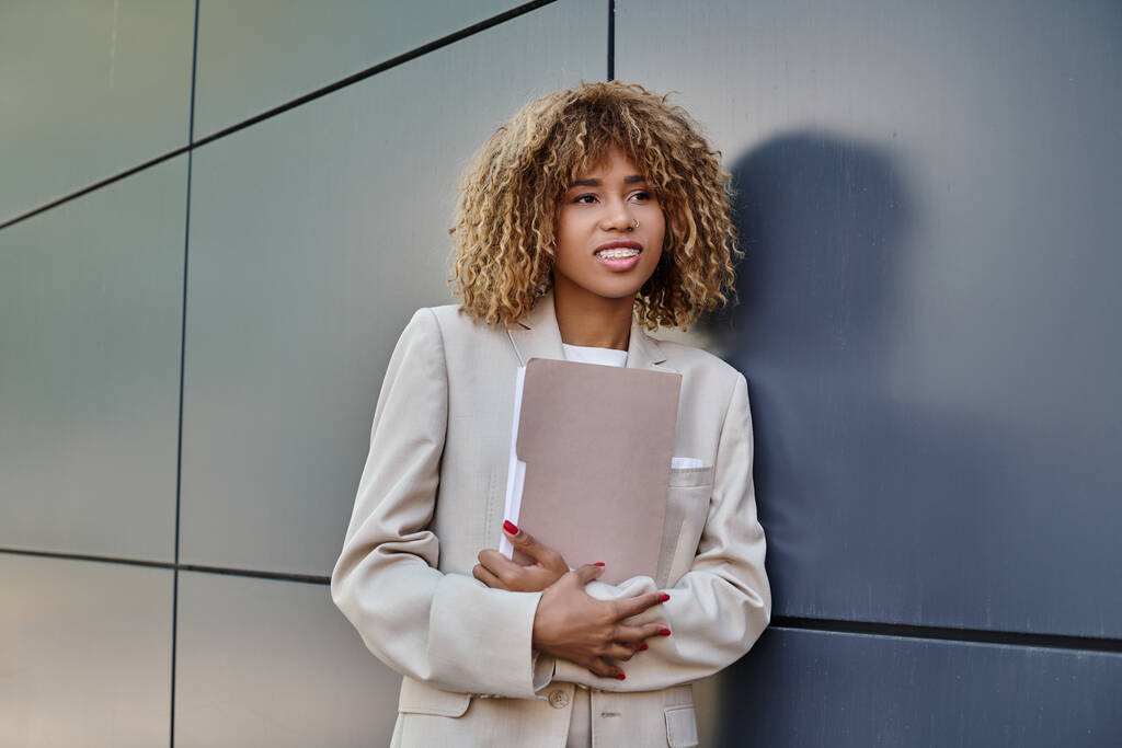 весела молода афроамериканська бізнес-леді в формальному носінні тримає папку сірою офісною стіною - Фото, зображення