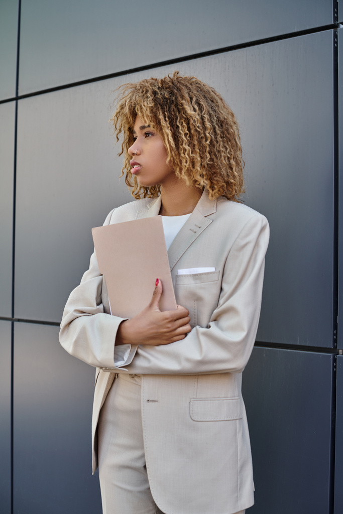 кудрявая и молодая африканская американская бизнесвумен в формальной одежде держит папку с серой офисной стеной - Фото, изображение