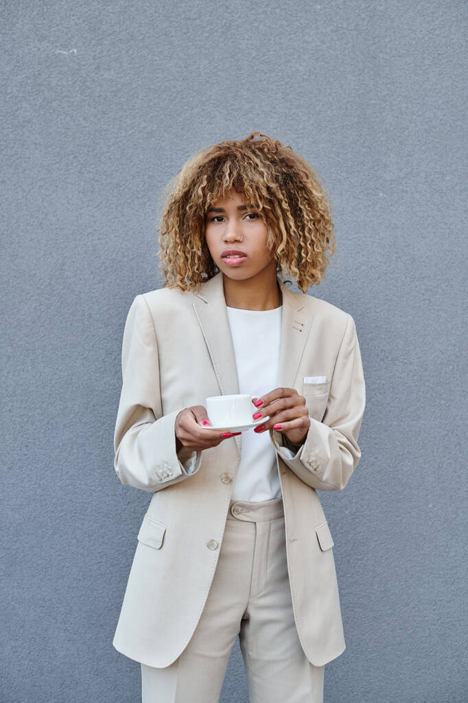 femme d'affaires afro-américaine frisée posant avec une tasse de café près du mur gris de l'immeuble de bureaux - Photo, image