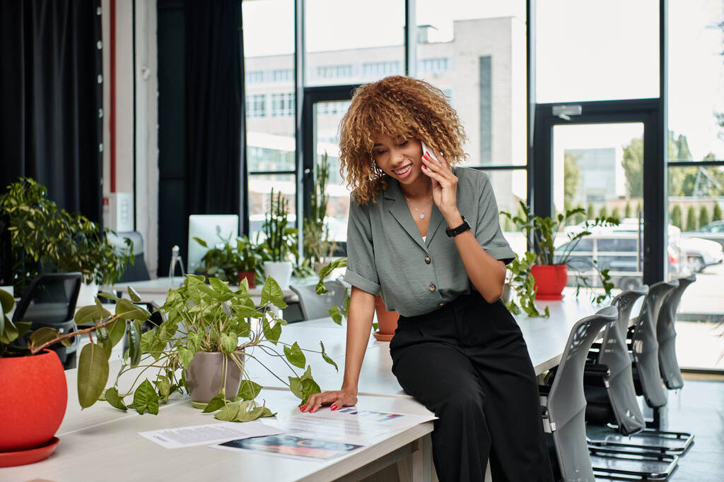 Empresária americana africana alegre durante telefonema olhando para gráficos na mesa de escritório - Foto, Imagem