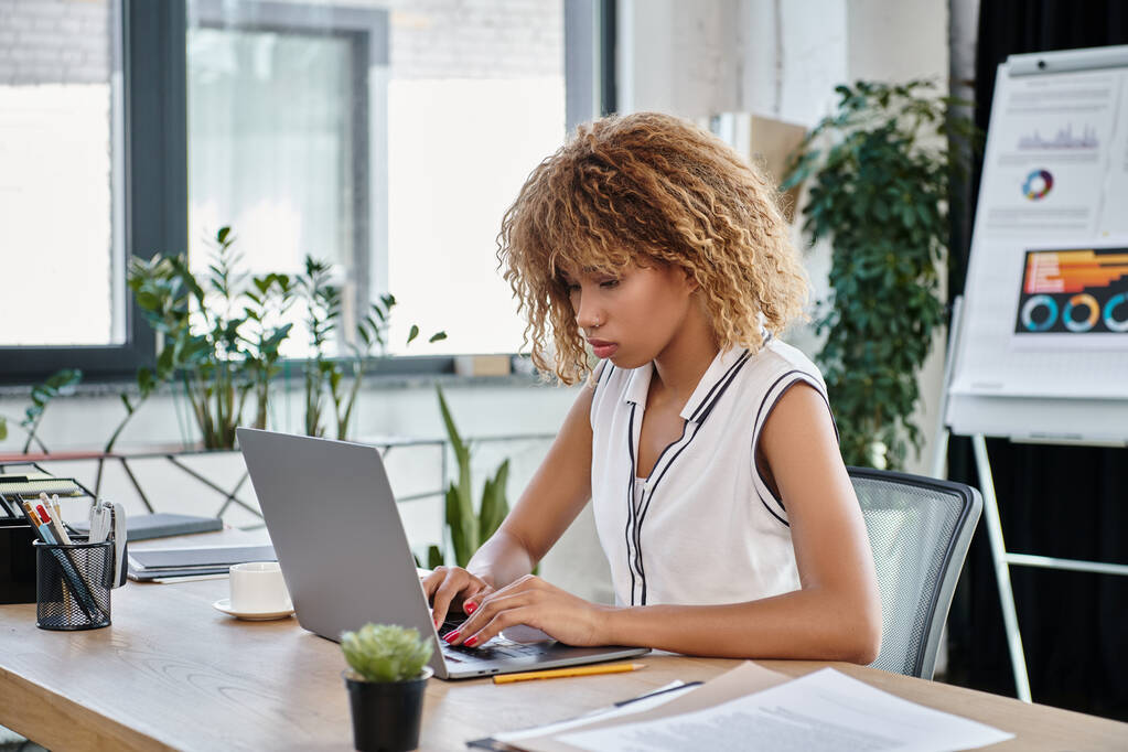 Kıvırcık Afrikalı Amerikan iş kadını, gündelik giysiler içinde, modern ofiste dizüstü bilgisayarda çalışıyor. - Fotoğraf, Görsel
