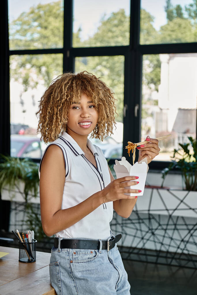 feliz encaracolado Africano americano mulher segurando takeaway refeição na caixa de papelão e olhando para a câmera - Foto, Imagem