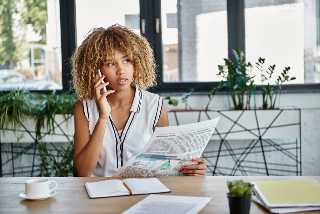 lockige afrikanisch-amerikanische Geschäftsfrau spricht mit Smartphone und hält Zeitung am Schreibtisch im Büro - Foto, Bild