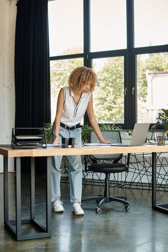сфокусированная молодая африканская американка, стоящая за своим столом и глядящая на ноутбук в течение рабочего дня - Фото, изображение