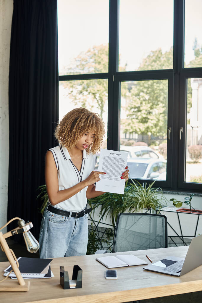 jeune femme afro-américaine debout à son bureau et montrant contrat lors d'un appel vidéo sur ordinateur portable - Photo, image